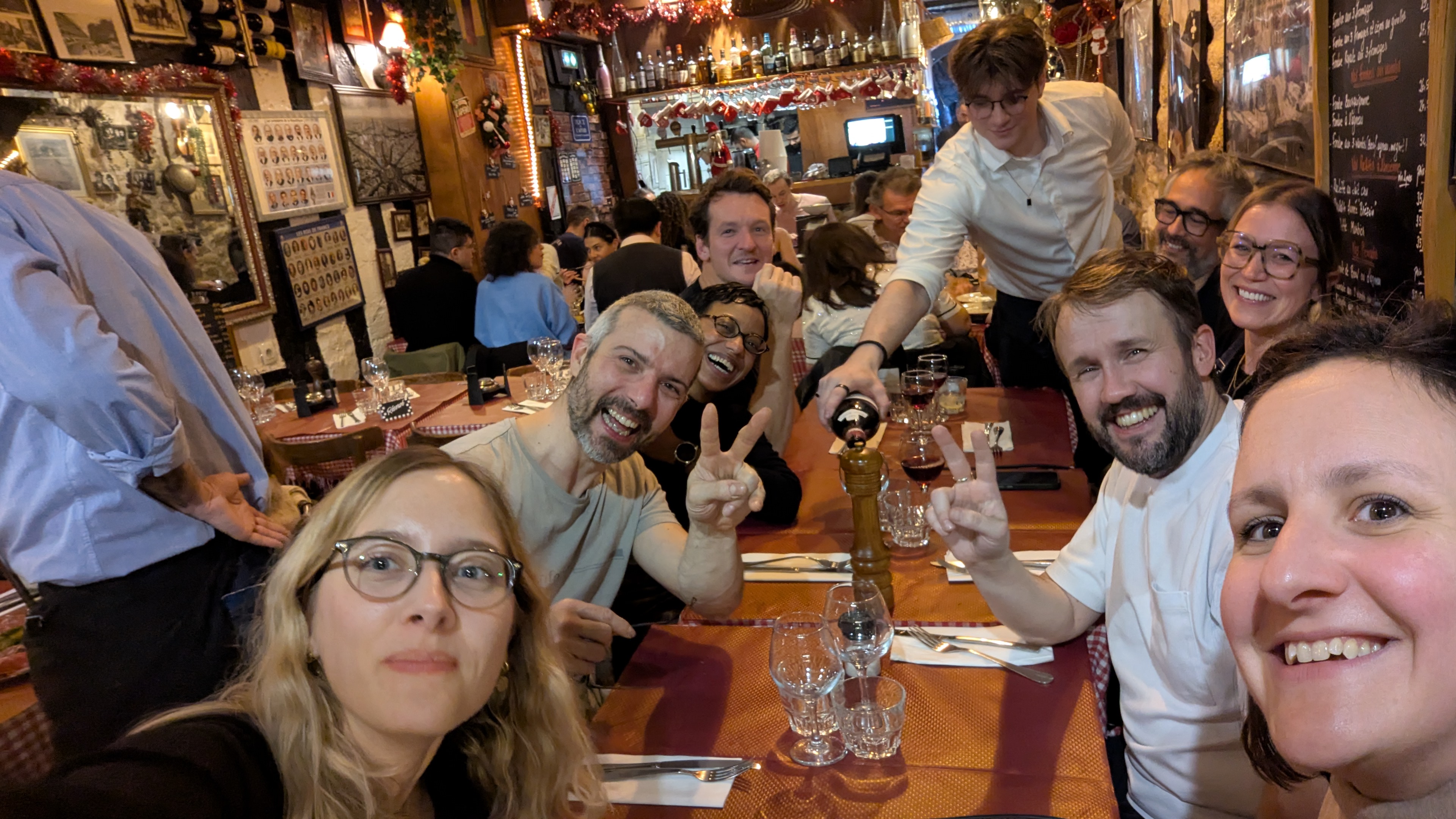 Selfie d’un groupe d’amis attablés dans un restaurant chaleureux, un serveur versant du vin.