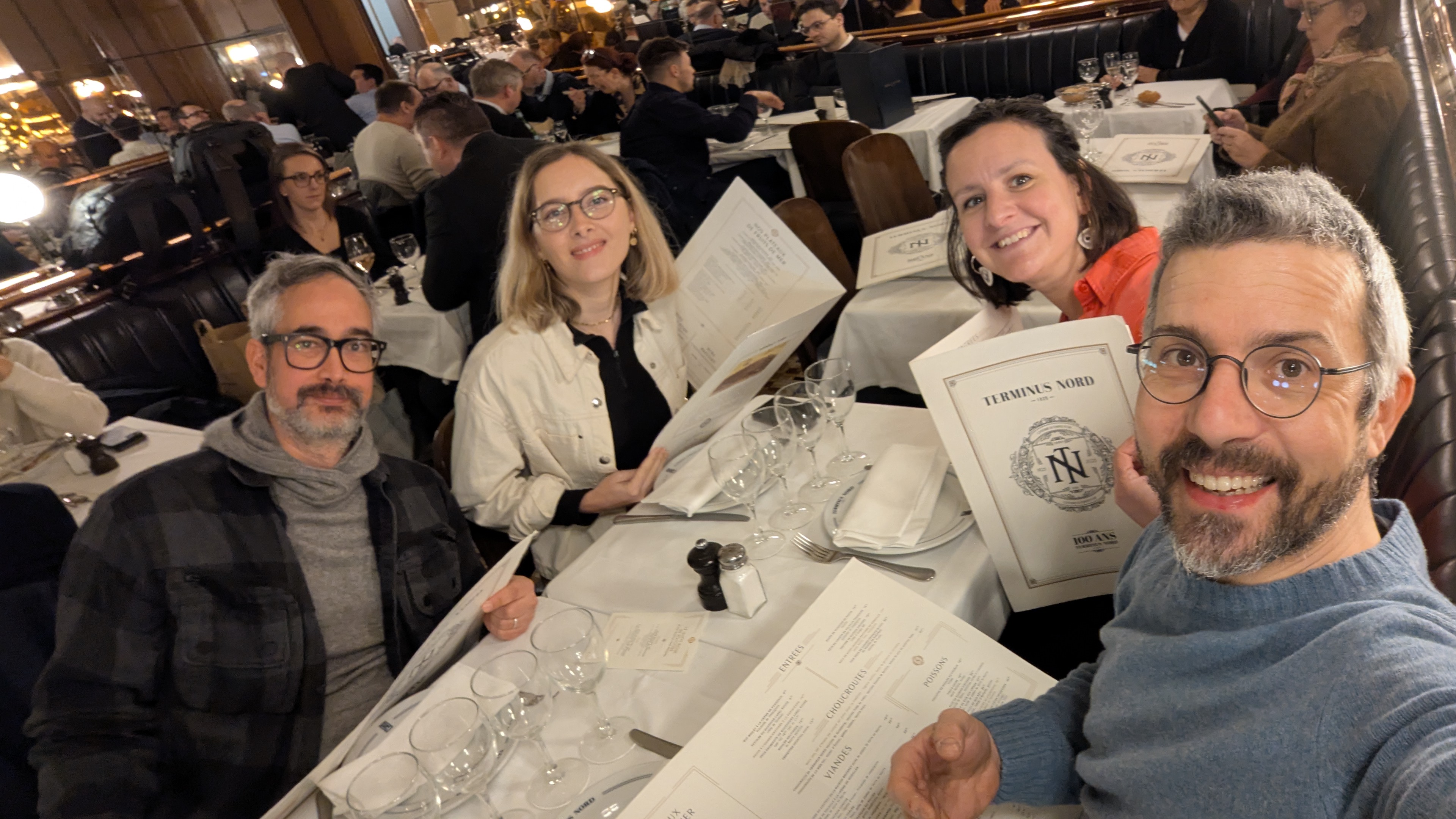 Quatre personnes attablées dans un restaurant parisien, tenant des menus “Terminus Nord” face à l’appareil photo.