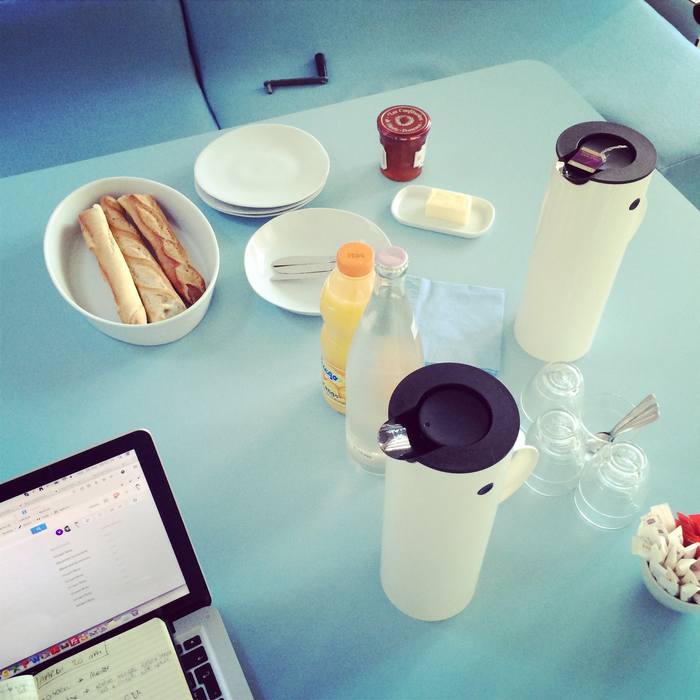 image d'une table de réunion avec un ordi sur la gauche et des baquettes avec beurre et confiture, des jus de fruits, une bouteille d'eau Evian, du café dans des thermos.