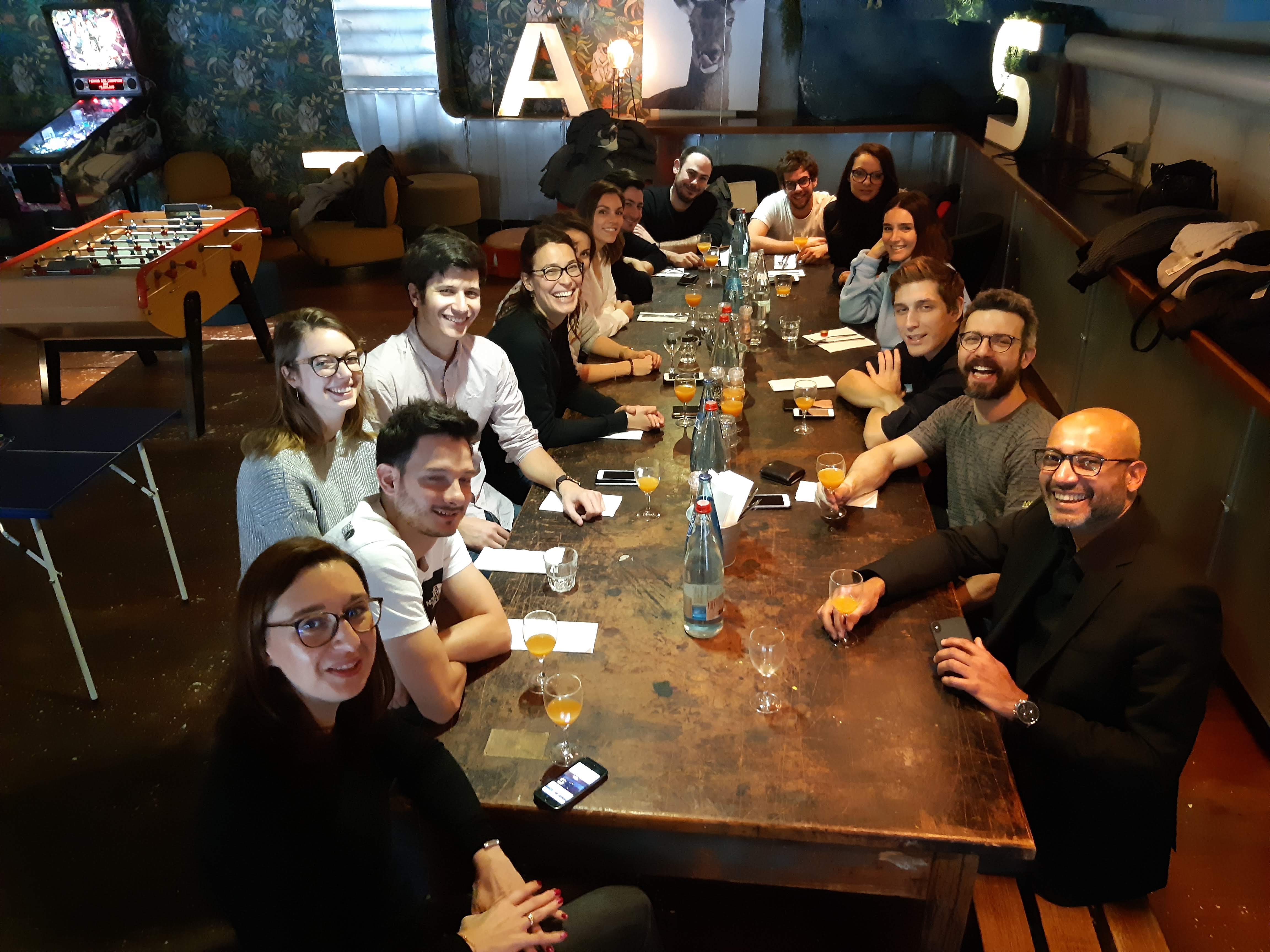 Groupe de collègues réunis autour d’une grande table en bois dans un bar, verres et bouteilles posés au centre.