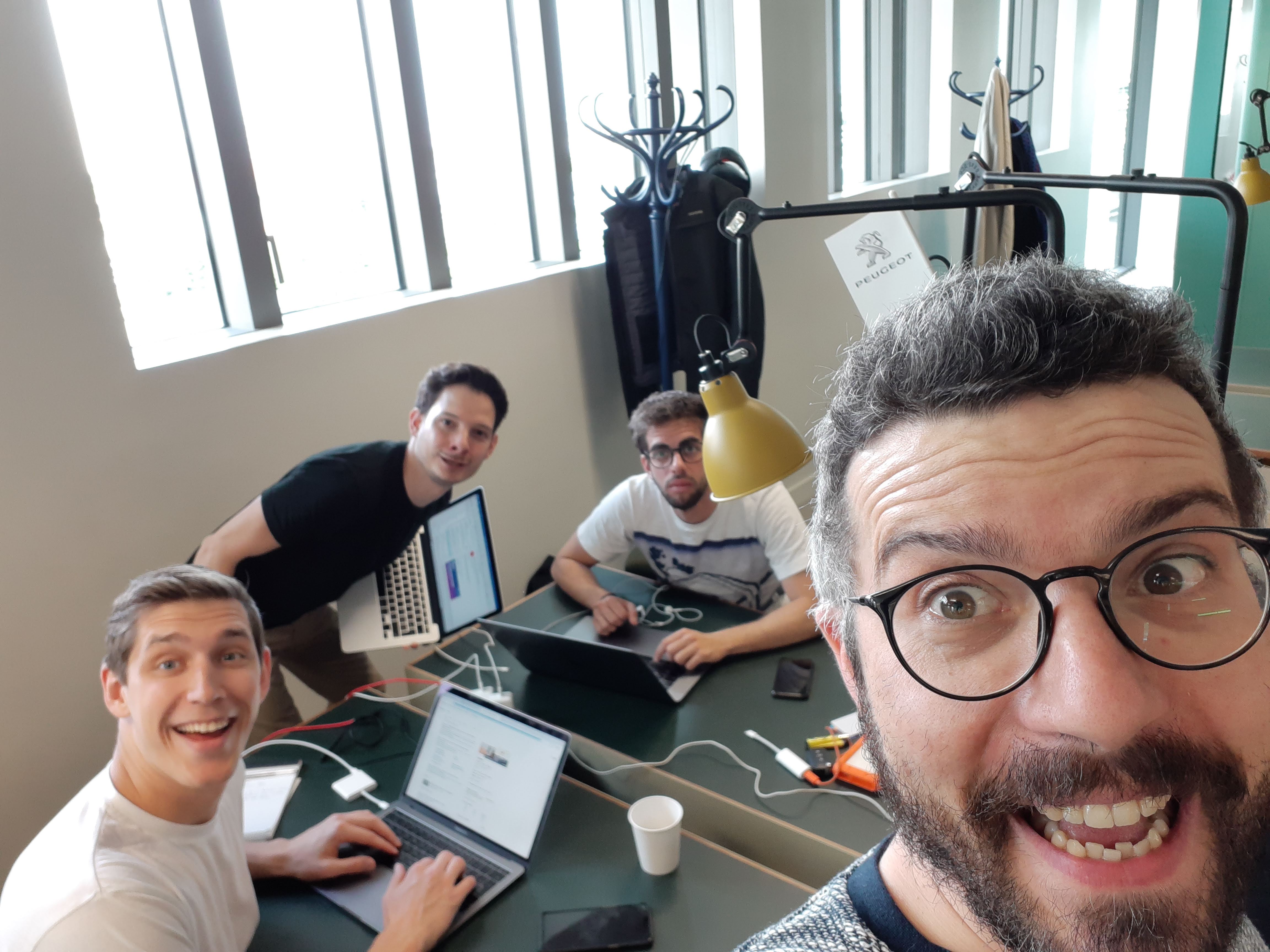 Quatre collègues travaillant sur ordinateurs portables autour d’une table de bureau, selfie pris au premier plan.