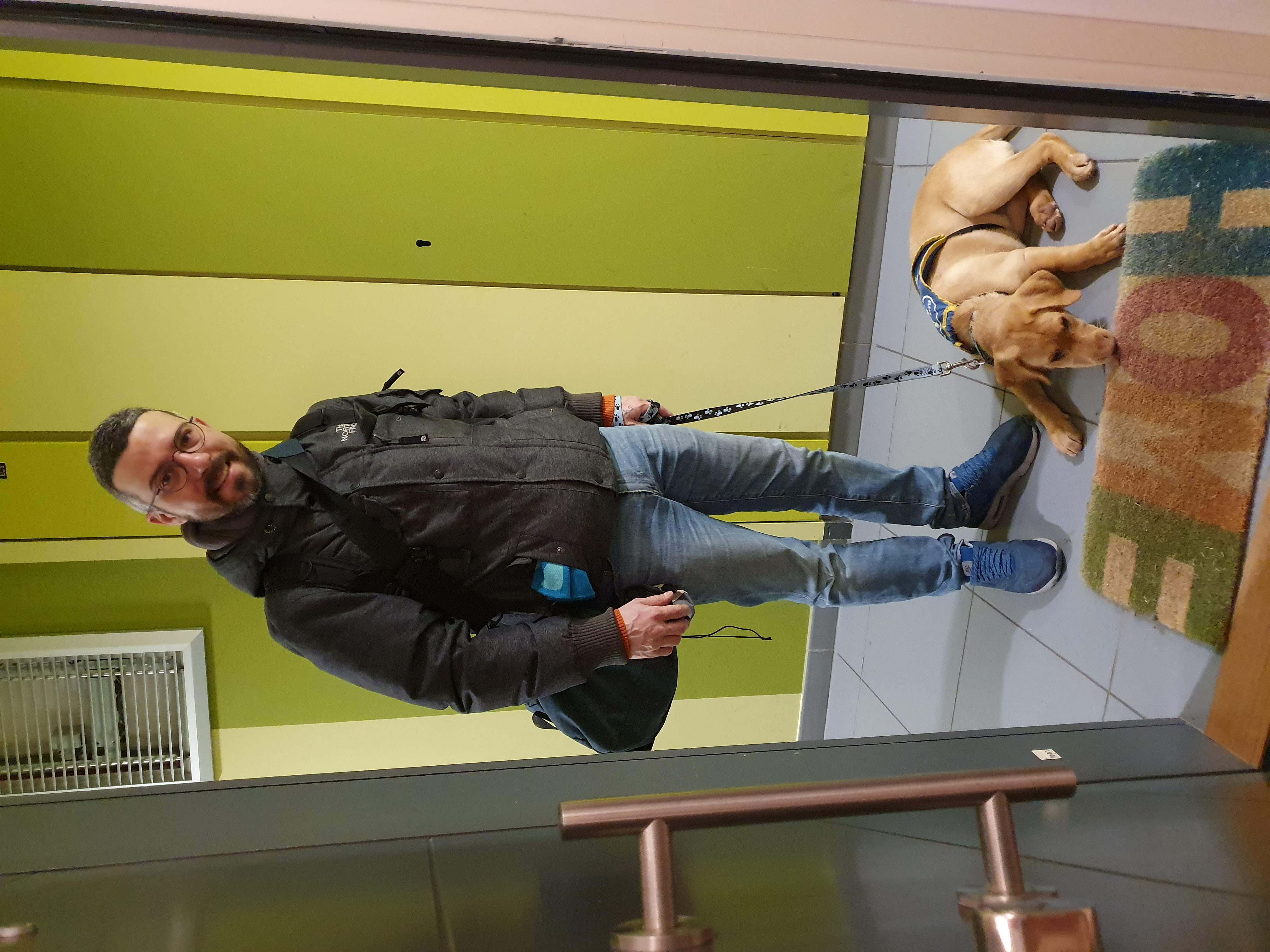 Homme tenant un chien en laisse dans l’entrée d’un bâtiment, le chien couché sur un paillasson coloré.