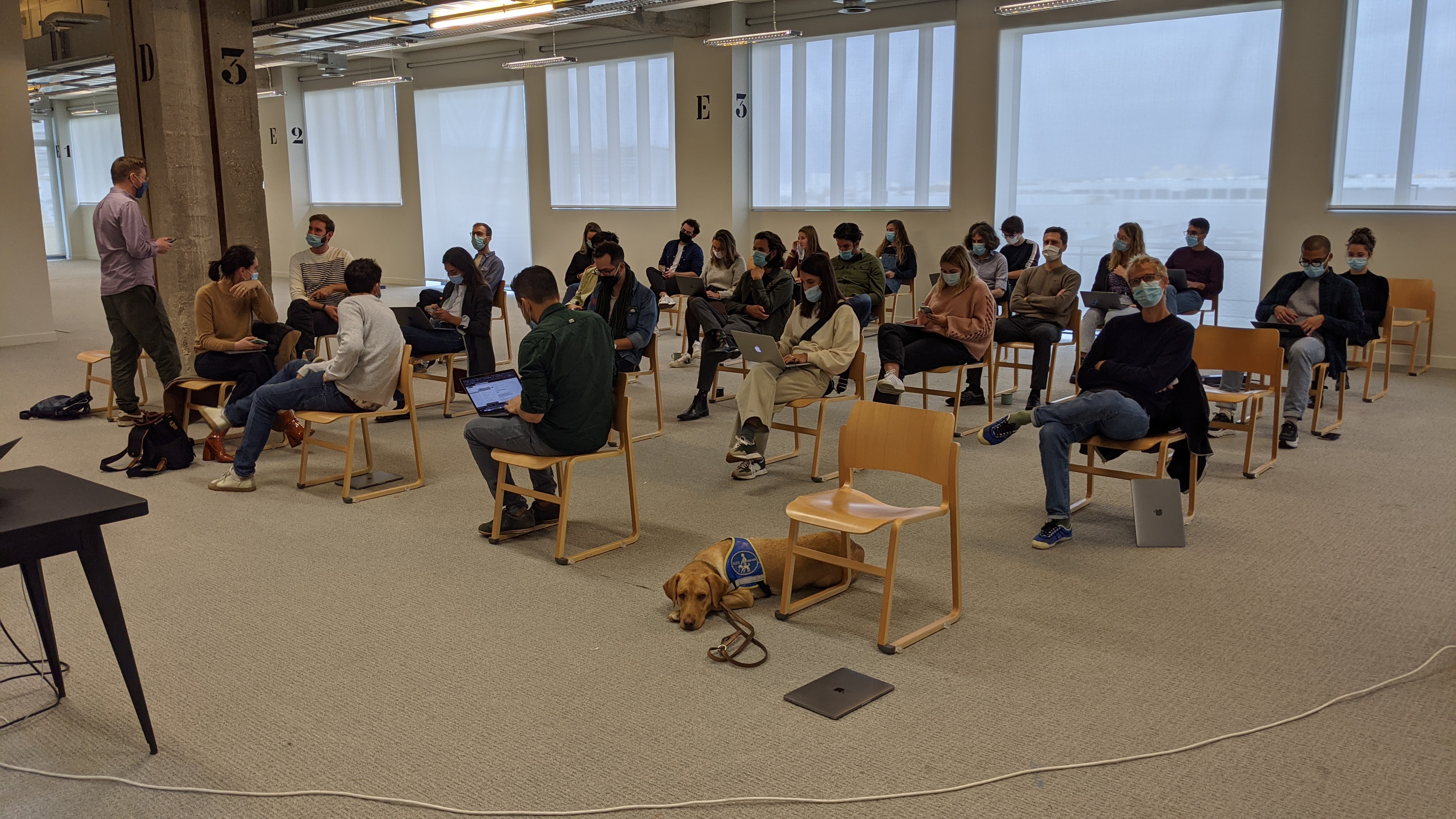 Conférence en salle avec public masqué assis sur des chaises espacées, chien guide allongé au centre.