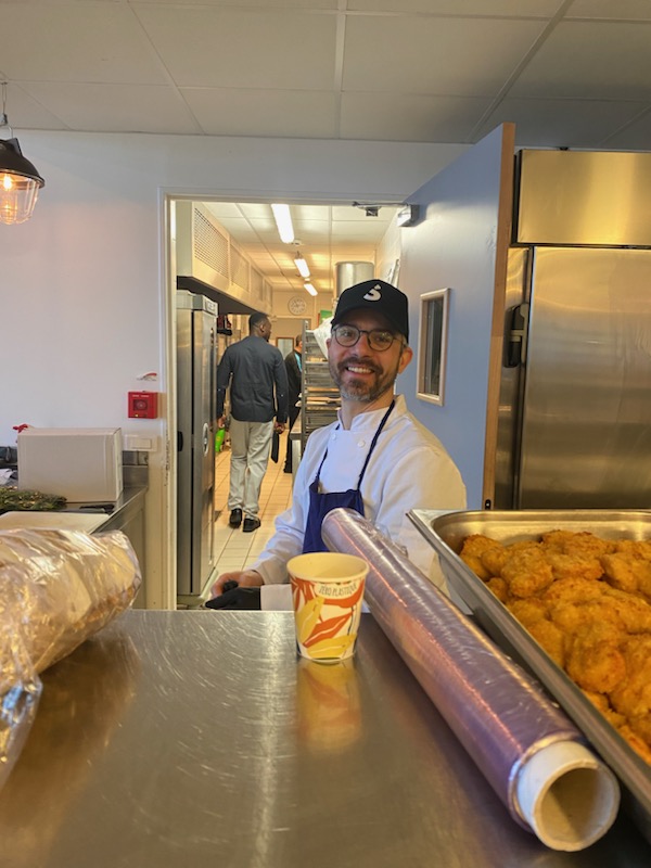 Homme en tenue de cuisine souriant derrière un comptoir, cuisine visible en arrière-plan.