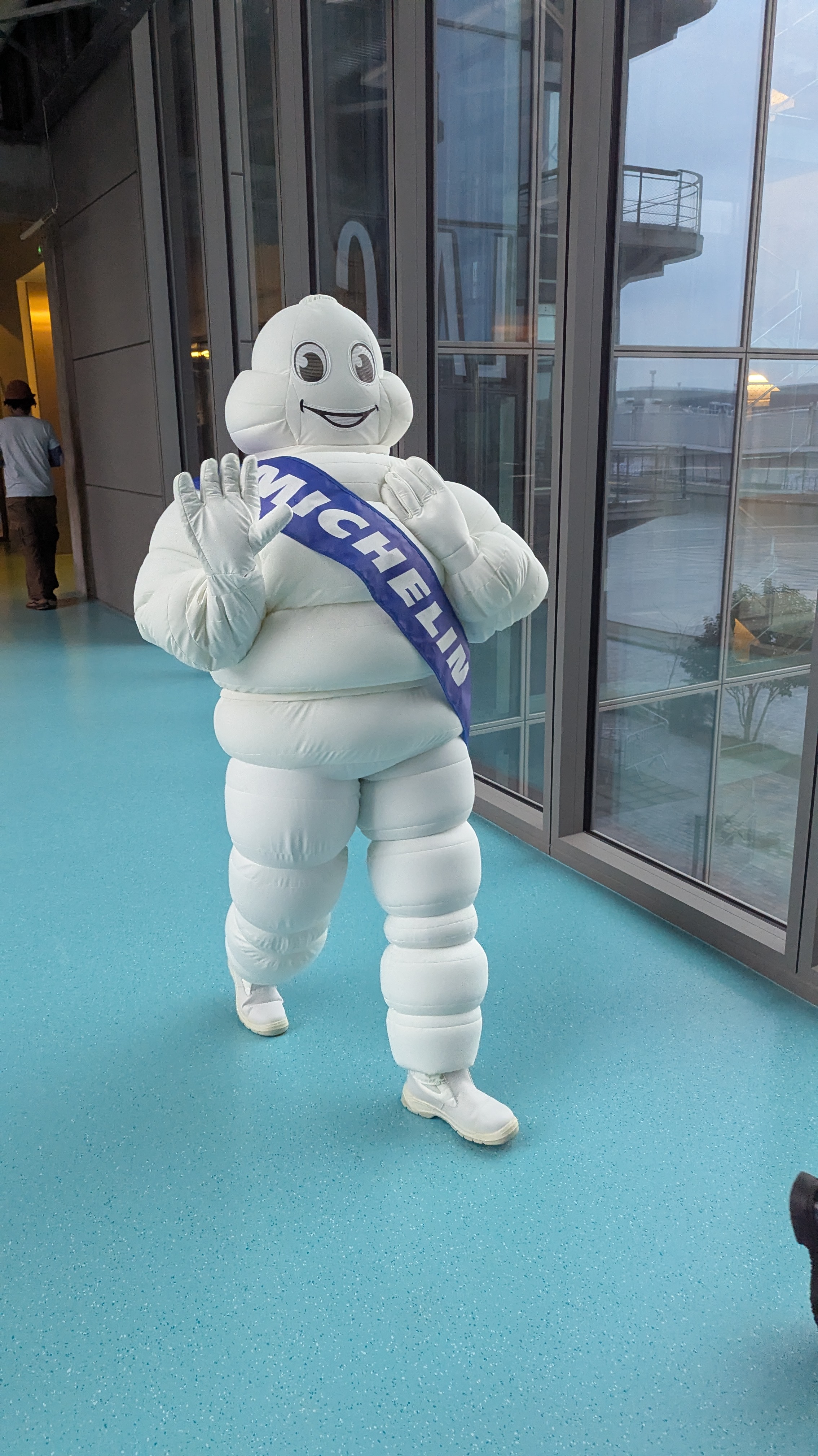 Mascotte Michelin en costume blanc posant dans un couloir vitré d’entreprise.